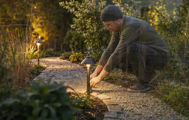 Person richtet eine niedrige warm leuchtende Gartenlampe am Kiesweg aus; Thema Pizzaöfen, Schwerpunkt Checkliste; Praxisalltag.