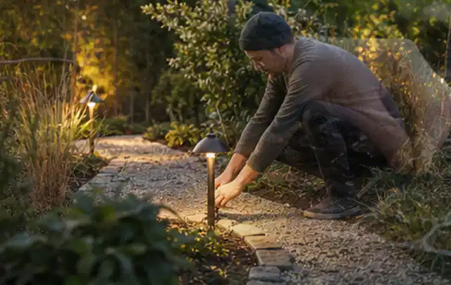 Person richtet eine niedrige warm leuchtende Gartenlampe am Kiesweg aus; Thema Pizzaöfen, Schwerpunkt Gärten mit kleiner Fläche; Praxisalltag.