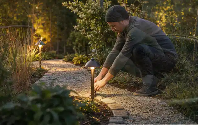 Person richtet eine niedrige warm leuchtende Gartenlampe am Kiesweg aus; Thema Pizzaöfen, Schwerpunkt Kaufberatung; Ergebnis.
