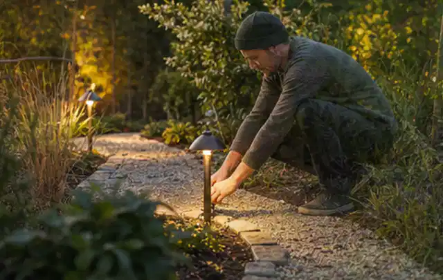 Person richtet eine niedrige warm leuchtende Gartenlampe am Kiesweg aus; Thema Pizzaöfen, Schwerpunkt Wartung; Kontrolle.
