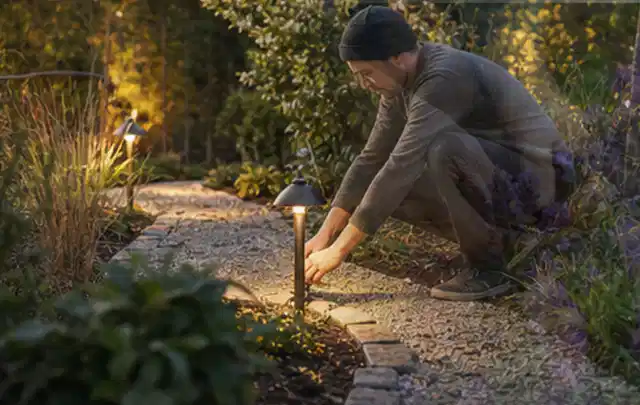 Person richtet eine niedrige warm leuchtende Gartenlampe am Kiesweg aus; Thema Poolabdeckungen, Schwerpunkt Anwendung; Kontrolle.