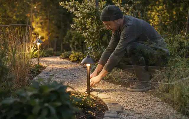 Person richtet eine niedrige warm leuchtende Gartenlampe am Kiesweg aus; Thema Poolabdeckungen, Schwerpunkt Bestenliste; Ausgangslage.
