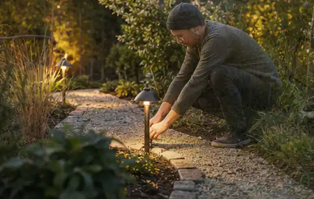 Person richtet eine niedrige warm leuchtende Gartenlampe am Kiesweg aus; Thema Poolabdeckungen, Schwerpunkt Checkliste; Umsetzung.