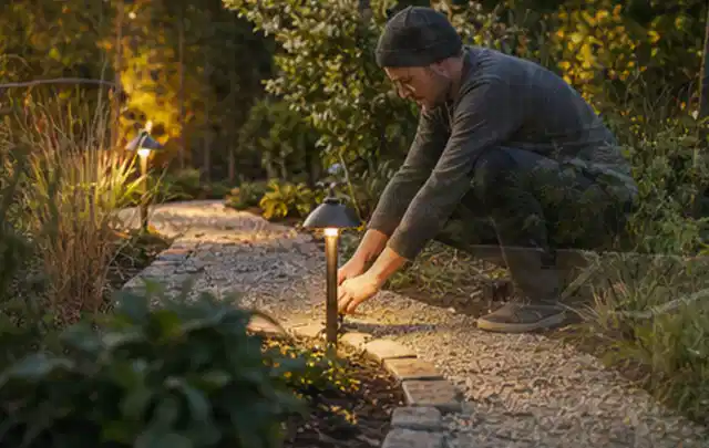 Person richtet eine niedrige warm leuchtende Gartenlampe am Kiesweg aus; Thema Poolabdeckungen, Schwerpunkt Kosten; Ausgangslage.
