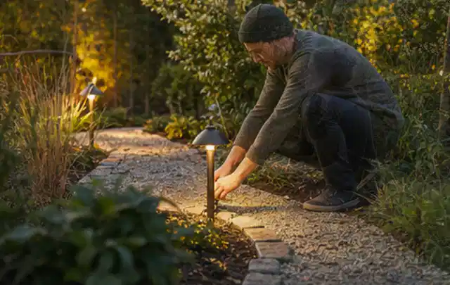 Person richtet eine niedrige warm leuchtende Gartenlampe am Kiesweg aus; Thema Poolabdeckungen, Schwerpunkt Testkriterien; Ergebnis.