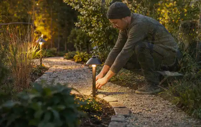 Person richtet eine niedrige warm leuchtende Gartenlampe am Kiesweg aus; Thema Poolabdeckungen, Schwerpunkt Winterlager; Ergebnis.