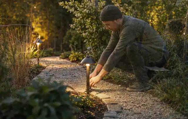 Person richtet eine niedrige warm leuchtende Gartenlampe am Kiesweg aus; Thema Poolchemie, Schwerpunkt Kosten; Kontrolle.