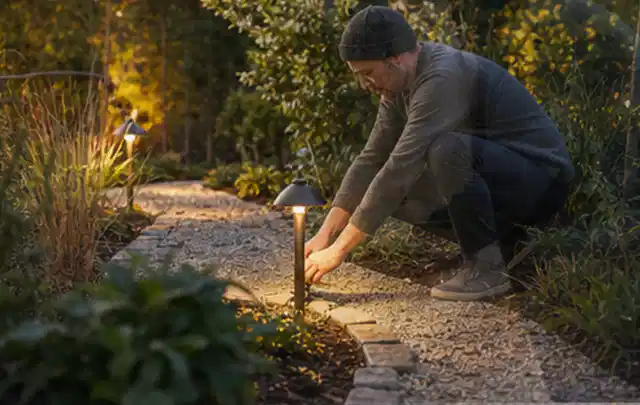 Person richtet eine niedrige warm leuchtende Gartenlampe am Kiesweg aus; Thema Poolchemie, Schwerpunkt Nachhaltigkeit; Umsetzung.