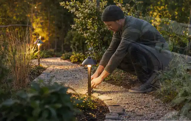 Person richtet eine niedrige warm leuchtende Gartenlampe am Kiesweg aus; Thema Poolchemie, Schwerpunkt Sicherheit; Praxisalltag.