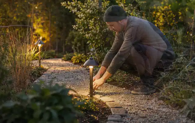Person richtet eine niedrige warm leuchtende Gartenlampe am Kiesweg aus; Thema Poolchemie, Schwerpunkt Systemvergleich; Kontrolle.