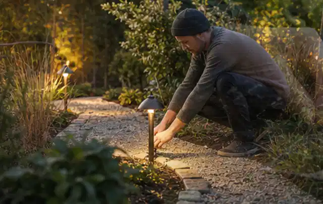 Person richtet eine niedrige warm leuchtende Gartenlampe am Kiesweg aus; Thema Poolchemie, Schwerpunkt Vergleich; Ergebnis.