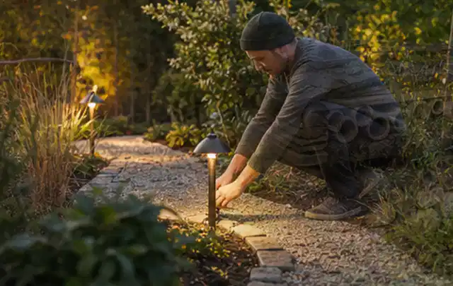 Person richtet eine niedrige warm leuchtende Gartenlampe am Kiesweg aus; Thema Poolchemie, Schwerpunkt Winterlager; Kontrolle.