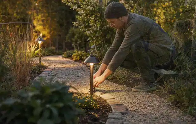 Person richtet eine niedrige warm leuchtende Gartenlampe am Kiesweg aus; Thema Poolfilter, Schwerpunkt Anwendung; Kontrolle.