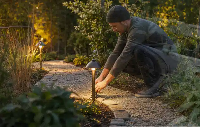 Person richtet eine niedrige warm leuchtende Gartenlampe am Kiesweg aus; Thema Poolfilter, Schwerpunkt Ersatzteile; Praxisalltag.