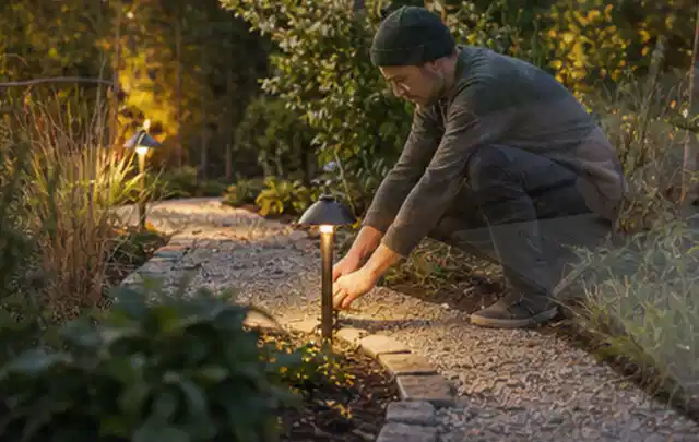 Person richtet eine niedrige warm leuchtende Gartenlampe am Kiesweg aus; Thema Poolfilter, Schwerpunkt Gärten mit großer Fläche; Kontrolle.