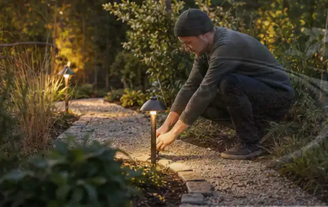 Person richtet eine niedrige warm leuchtende Gartenlampe am Kiesweg aus; Thema Poolfilter, Schwerpunkt Gärten mit kleiner Fläche; Umsetzung.