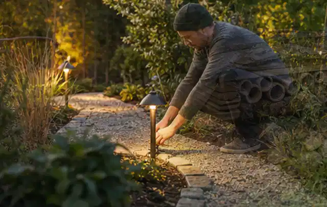 Person richtet eine niedrige warm leuchtende Gartenlampe am Kiesweg aus; Thema Poolfilter, Schwerpunkt Sicherheit; Kontrolle.