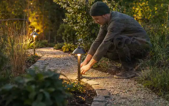 Person richtet eine niedrige warm leuchtende Gartenlampe am Kiesweg aus; Thema Poolfilter, Schwerpunkt Winterlager; Ausgangslage.