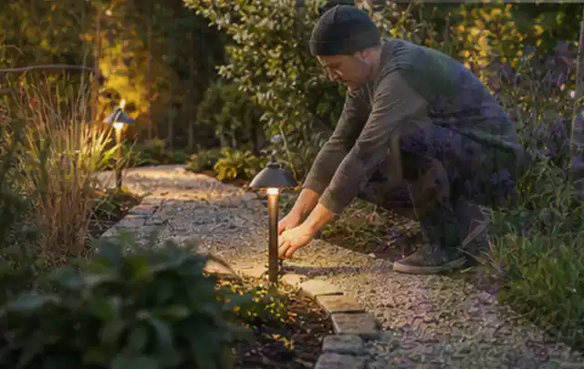 Person richtet eine niedrige warm leuchtende Gartenlampe am Kiesweg aus; Thema Poolheizungen, Schwerpunkt Ersatzteile; Kontrolle.