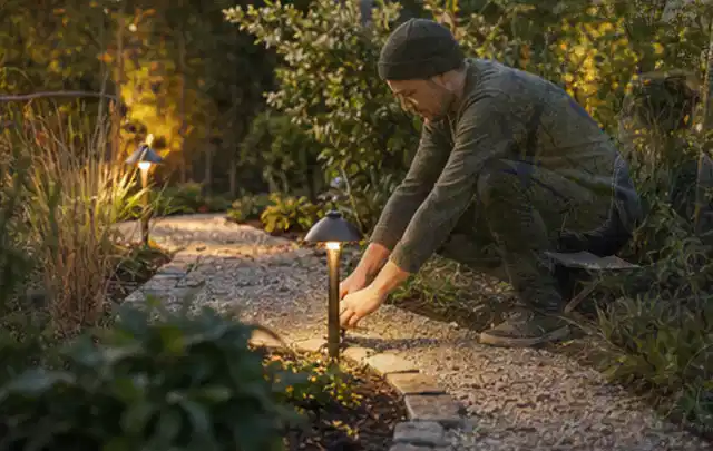 Person richtet eine niedrige warm leuchtende Gartenlampe am Kiesweg aus; Thema Poolheizungen, Schwerpunkt Nachhaltigkeit; Ergebnis.