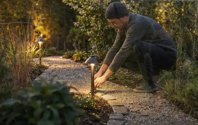 Person richtet eine niedrige warm leuchtende Gartenlampe am Kiesweg aus; Thema Poolheizungen, Schwerpunkt Wartung; Umsetzung.
