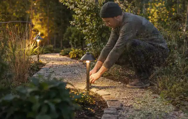 Person richtet eine niedrige warm leuchtende Gartenlampe am Kiesweg aus; Thema Poolheizungen, Schwerpunkt Winterlager; Ausgangslage.