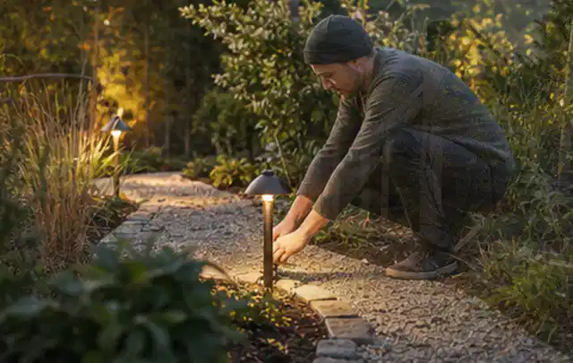Person richtet eine niedrige warm leuchtende Gartenlampe am Kiesweg aus; Thema Poolpflege, Schwerpunkt Testkriterien; Praxisalltag.