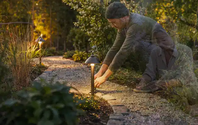 Person richtet eine niedrige warm leuchtende Gartenlampe am Kiesweg aus; Thema Poolpflege, Schwerpunkt Vergleich; Umsetzung.