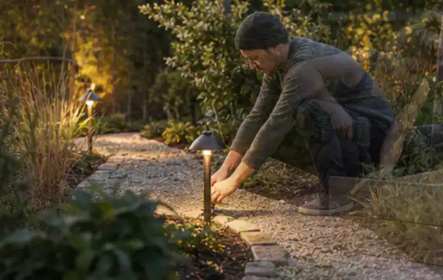 Person richtet eine niedrige warm leuchtende Gartenlampe am Kiesweg aus; Thema Poolpumpen, Schwerpunkt Checkliste; Kontrolle.