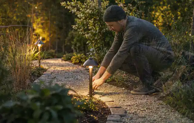 Person richtet eine niedrige warm leuchtende Gartenlampe am Kiesweg aus; Thema Poolpumpen, Schwerpunkt Systemvergleich; Kontrolle.