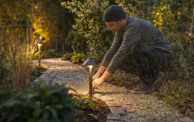 Person richtet eine niedrige warm leuchtende Gartenlampe am Kiesweg aus; Thema Poolpumpen, Schwerpunkt Vergleich; Ergebnis.