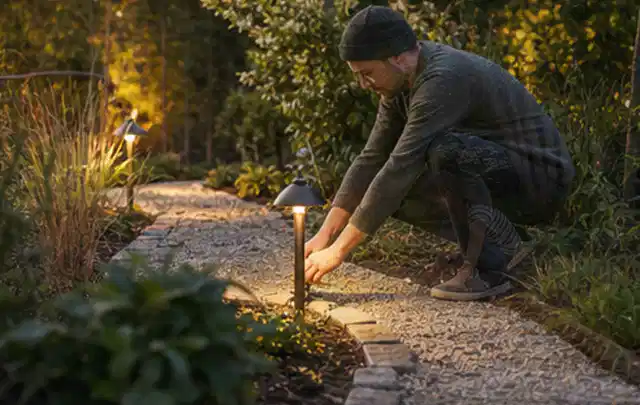 Person richtet eine niedrige warm leuchtende Gartenlampe am Kiesweg aus; Thema Poolroboter, Schwerpunkt Checkliste; Praxisalltag.