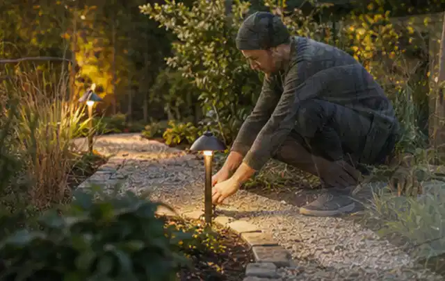Person richtet eine niedrige warm leuchtende Gartenlampe am Kiesweg aus; Thema Poolroboter, Schwerpunkt Dimensionierung; Ergebnis.
