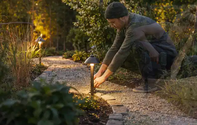 Person richtet eine niedrige warm leuchtende Gartenlampe am Kiesweg aus; Thema Poolroboter, Schwerpunkt Fehlerhilfe; Praxisalltag.