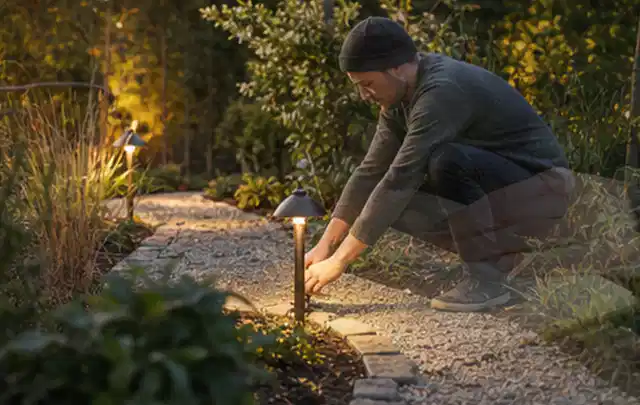 Person richtet eine niedrige warm leuchtende Gartenlampe am Kiesweg aus; Thema Poolroboter, Schwerpunkt Pools; Ergebnis.