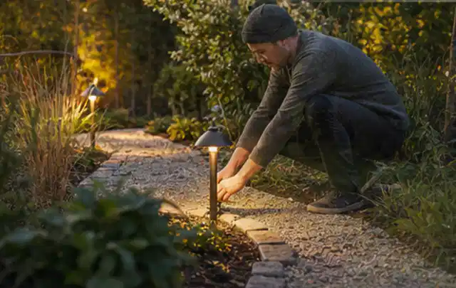 Person richtet eine niedrige warm leuchtende Gartenlampe am Kiesweg aus; Thema Poolroboter, Schwerpunkt Reparatur; Ausgangslage.