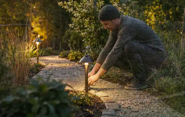 Person richtet eine niedrige warm leuchtende Gartenlampe am Kiesweg aus; Thema Poolroboter, Schwerpunkt Sicherheit; Kontrolle.