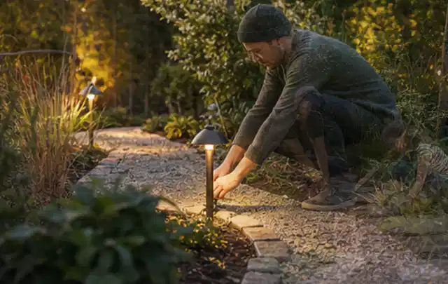 Person richtet eine niedrige warm leuchtende Gartenlampe am Kiesweg aus; Thema Poolroboter, Schwerpunkt Systemvergleich; Kontrolle.