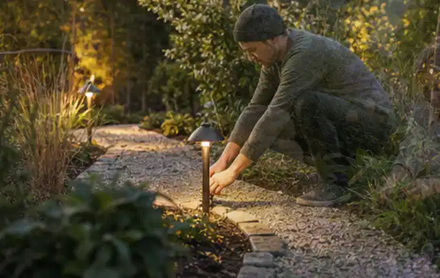 Person richtet eine niedrige warm leuchtende Gartenlampe am Kiesweg aus; Thema Poolroboter, Schwerpunkt Testkriterien; Praxisalltag.