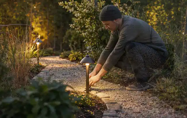 Person richtet eine niedrige warm leuchtende Gartenlampe am Kiesweg aus; Thema Rankpflanzen als Sichtschutz, Schwerpunkt Baurecht; Praxisalltag.