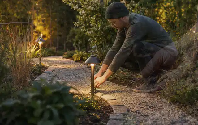 Person richtet eine niedrige warm leuchtende Gartenlampe am Kiesweg aus; Thema Rankpflanzen als Sichtschutz, Schwerpunkt Checkliste; Umsetzung.