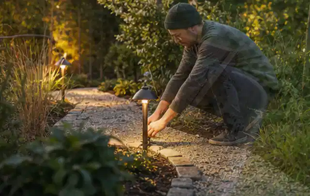 Person richtet eine niedrige warm leuchtende Gartenlampe am Kiesweg aus; Thema Rankpflanzen als Sichtschutz, Schwerpunkt Fundament; Umsetzung.