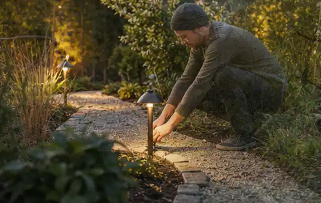 Person richtet eine niedrige warm leuchtende Gartenlampe am Kiesweg aus; Thema Rankpflanzen als Sichtschutz, Schwerpunkt Gärten mit kleiner Fläche;…