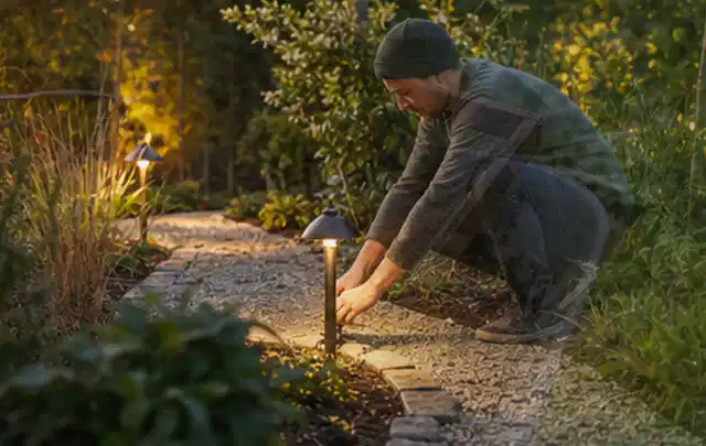 Person richtet eine niedrige warm leuchtende Gartenlampe am Kiesweg aus; Thema Rankpflanzen als Sichtschutz, Schwerpunkt Gestaltung; Ergebnis.
