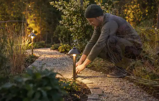 Person richtet eine niedrige warm leuchtende Gartenlampe am Kiesweg aus; Thema Rankpflanzen als Sichtschutz, Schwerpunkt Materialwahl; Kontrolle.