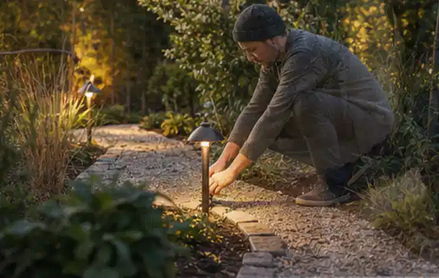 Person richtet eine niedrige warm leuchtende Gartenlampe am Kiesweg aus; Thema Rankpflanzen als Sichtschutz, Schwerpunkt Nachhaltigkeit; Ausgangslage.
