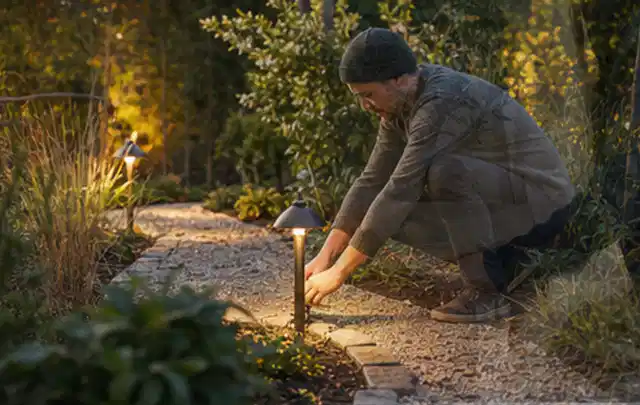 Person richtet eine niedrige warm leuchtende Gartenlampe am Kiesweg aus; Thema Rankpflanzen als Sichtschutz, Schwerpunkt Standort; Umsetzung.