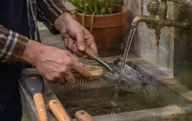 Hände reinigen gebrauchte Gartengeräte mit Bürste und Wasser am Außenbecken; Thema Rasenmäher, Schwerpunkt Ersatzteile; Ergebnis.