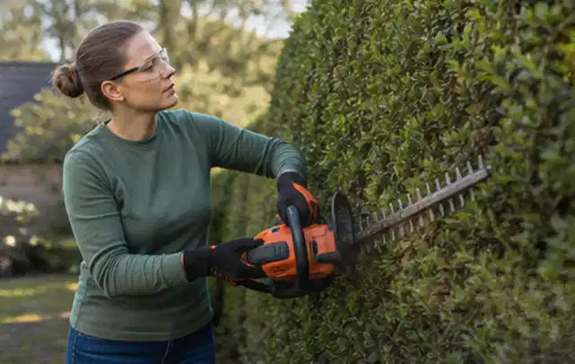 Person schneidet eine dichte Gartenhecke mit Schutzbrille und sicherem Stand; Thema Rasenmäher, Schwerpunkt Kaufberatung; Praxisalltag.