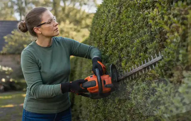 Person schneidet eine dichte Gartenhecke mit Schutzbrille und sicherem Stand; Thema Rasenmäher, Schwerpunkt Kosten; Praxisalltag.